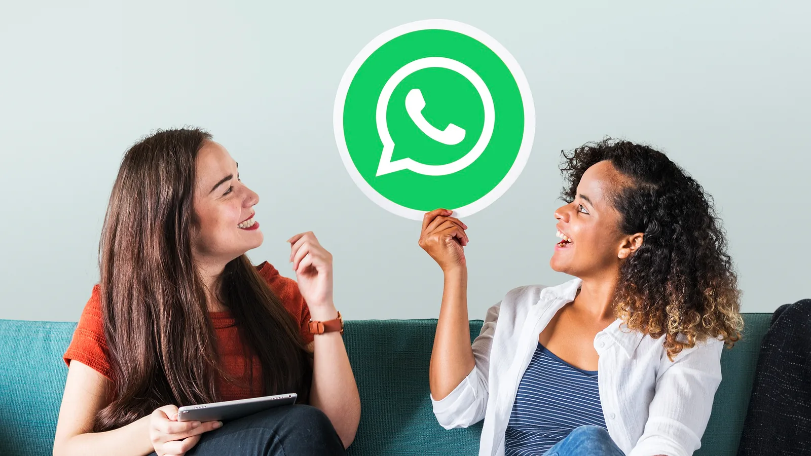 Two friends on a sofa laughing, one holding up a WhatsApp logo sign